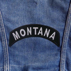 May include: A black, arched patch with the word "MONTANA" embroidered in white, sewn onto a blue denim jacket. The patch is centered and slightly curved, with the letters clearly visible against the dark background.