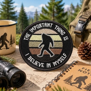 May include: A black embroidered patch with the text "THE IMPORTANT THING IS I BELIEVE IN MYSELF" and a Bigfoot silhouette. The patch is round with a striped background of beige, green, and brown. A beige enamel mug with a Bigfoot graphic is in the background.