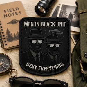 May include: A rectangular black embroidered patch featuring two figures in suits, hats, and sunglasses. The text reads MEN IN BLACK UNIT at the top and DENY EVERYTHING at the bottom. A small flying saucer graphic is positioned above the figures.