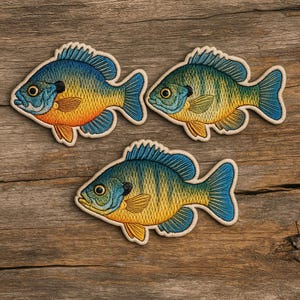 May include: Three embroidered patches in the shape of bluegill fish. The patches feature blue, yellow, and orange colors with detailed stitching. The patches are set against a wooden background.