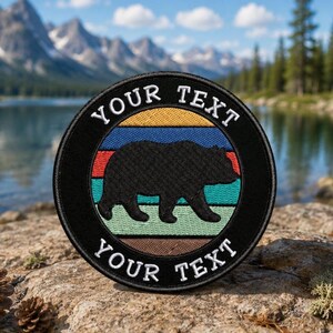 May include: A circular embroidered patch featuring a black bear silhouette centered over horizontal stripes of yellow, blue, red, and green. The black border contains the white embroidered text YOUR TEXT at the top and bottom.