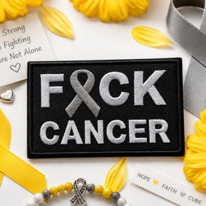 Może przedstawiać: Prostokątna czarna naszywka z wyhaftowanym srebrnym napisem F-wstążka-CK CANCER. Litera O została zastąpiona srebrną wstążką świadomości. Naszywka jest otoczona żółtymi płatkami kwiatów, szarą wstążką oraz białą kartką z nadrukowanym tekstem.