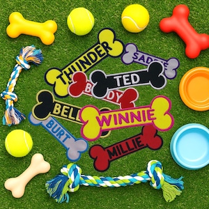 May include: A collection of dog toys and accessories on green grass. Includes yellow and red bone-shaped toys, tennis balls, rope toys, and personalized bone-shaped patches with names like "Thunder" and "Winnie". An orange and a blue bowl are also present.