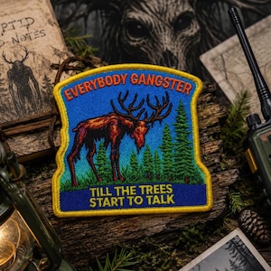 May include: A shield-shaped embroidered patch with a blue background, featuring a brown moose with large antlers standing in a green forest. The text reads EVERYBODY GANGSTER at the top and TILL THE TREES START TO TALK at the bottom in yellow.