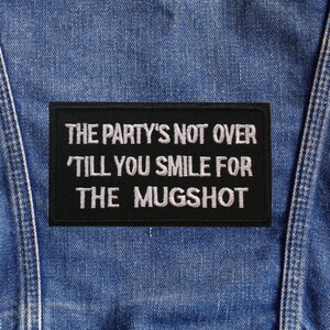 May include: A black rectangular patch with white text that reads "THE PARTY'S NOT OVER 'TILL YOU SMILE FOR THE MUGSHOT." The patch is sewn onto a blue denim fabric, likely a jacket or vest.