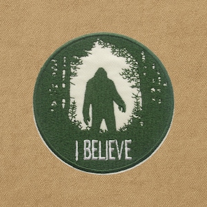 May include: A round, green embroidered patch featuring a silhouette of Bigfoot in a forest scene. The patch has a white border and the words "I BELIEVE" are embroidered in white at the bottom. The background is a tan fabric.