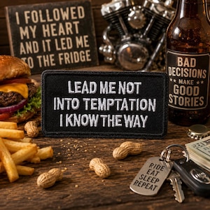 May include: A rectangular black embroidered patch featuring the white text "LEAD ME NOT INTO TEMPTATION I KNOW THE WAY" with a stitched border, positioned on a wooden surface alongside a burger, french fries, peanuts, and a beer bottle.