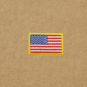 May include: A rectangular embroidered patch featuring the United States flag with a bright yellow border, stitched onto a tan, textured fabric background. The flag displays a blue canton with white stars and alternating red and white stripes.