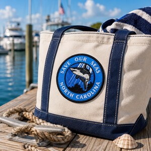 May include: A canvas tote bag with navy blue trim and handles. The bag features a circular patch with a shark graphic and the text "SAVE OUR SEAS NORTH CAROLINA". A striped towel is visible inside the bag.