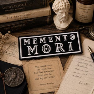 May include: A black patch with the words "MEMENTO MORI" embroidered in white. The patch is rectangular and sits on a wooden surface with books, a bust, and handwritten notes. The phrase is a reminder of mortality.