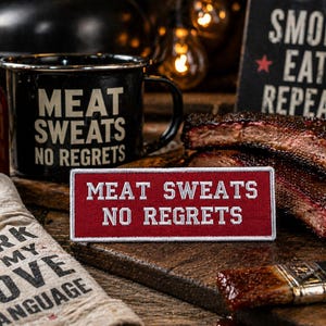 May include: A black enamel mug and a red patch with the text "MEAT SWEATS NO REGRETS" are displayed. A sign reads "SMOKE EAT REPEAT" next to cooked ribs. A bottle of sauce and a linen with text are also visible.