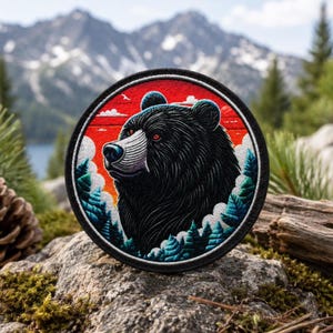 Puede incluir: Un parche bordado circular que presenta la cabeza de un oso negro de perfil. El fondo muestra un cielo rojo vibrante con nubes blancas sobre un bosque de pinos de color verde azulado. El parche tiene un borde negro grueso y costuras detalladas.