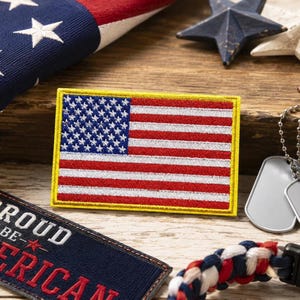 May include: A rectangular embroidered patch featuring the United States flag with a yellow border. The patch displays a blue field with white stars and alternating red and white horizontal stripes. The design is stitched with a textured finish.