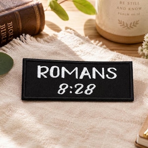 May include: A rectangular black embroidered patch featuring the white text ROMANS 8:28 in a bold, sans-serif font for the word ROMANS and a stylized script font for the numbers, with a finished stitched border surrounding the entire design.