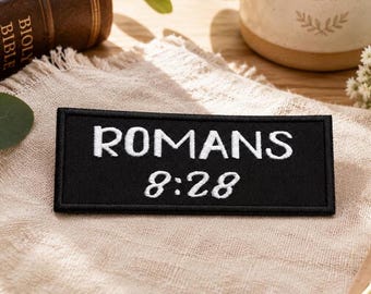 Op de afbeelding: Een rechthoekige zwarte geborduurde patch met de witte tekst ROMANS 8:28, waarbij het woord ROMANS in een vetgedrukt schreefloos lettertype staat en de cijfers in een gestileerd cursief lettertype, afgewerkt met een gestikte rand rondom.