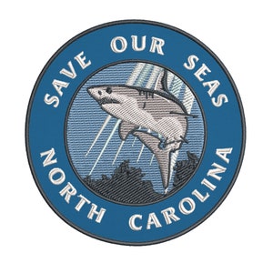 May include: Embroidered patch with a blue border and the text "SAVE OUR SEAS NORTH CAROLINA". The patch features a detailed illustration of a shark leaping from the water, with light blue rays and a dark gray landscape.