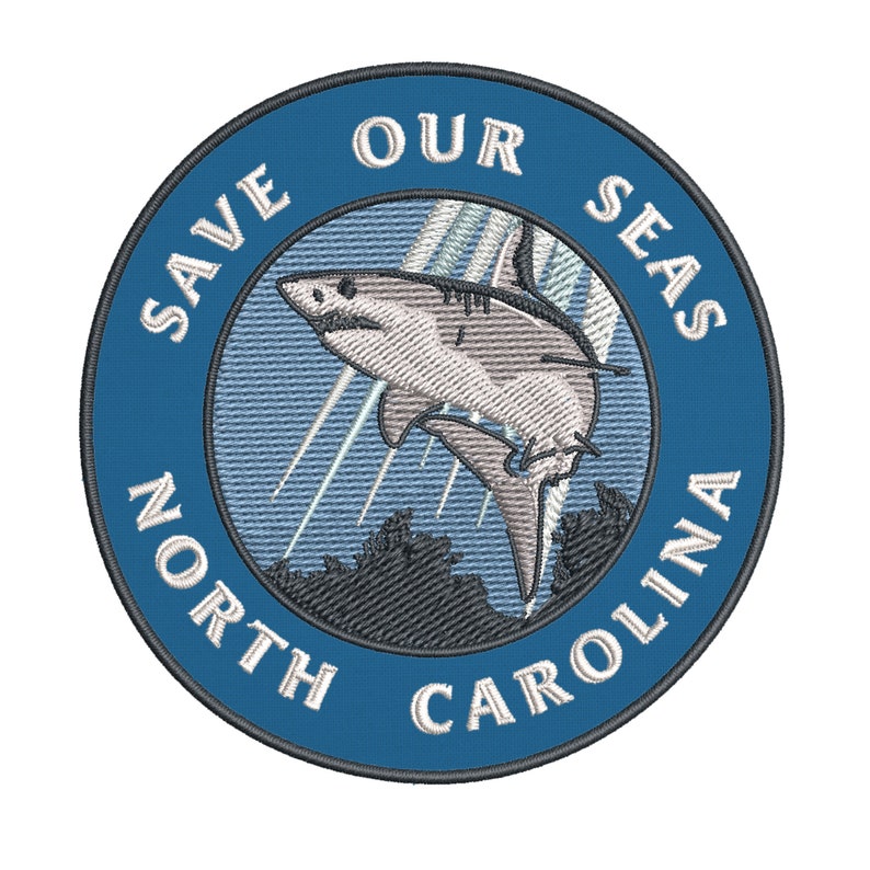 May include: Embroidered patch with a blue border and the text "SAVE OUR SEAS NORTH CAROLINA". The patch features a detailed illustration of a shark leaping from the water, with light blue rays and a dark gray landscape.
