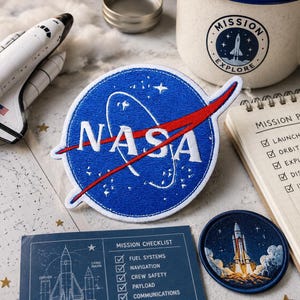 Könnte beinhalten: Ein runder, bestickter NASA-Aufnäher mit blauem Hintergrund, weißer Schrift und einem roten Bogen. Der Aufnäher ist umgeben von einem Space-Shuttle-Modell, einem Becher mit Missionslogo, einem Spiralblock und einer Missions-Checkliste.