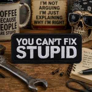 Puede incluir: Un parche rectangular negro bordado con texto blanco cosido que dice YOU CAN'T FIX STUPID. El parche presenta una fuente negrita y clara con un borde acabado, diseñado para su aplicación en artículos de tela como chaquetas, bolsos o gorras.