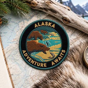 Könnte beinhalten: Runder, gestickter Aufnäher mit dem goldenen Schriftzug "ALASKA ADVENTURE AWAITS". Er zeigt braune Bären, türkisfarbenes Wasser und einen Fisch. Mit schwarzem Rand und türkisem Hintergrund.