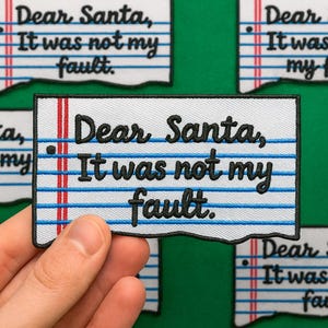 May include: Embroidered patch with the text "Dear Santa, It was not my fault" in black on a white background, resembling lined paper. The patch has a torn edge design, with red and blue lines. Several patches are visible.