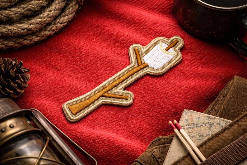 May include: Embroidered patch featuring a marshmallow on a stick, with a white marshmallow and a tan stick. The patch is on a red background with a pinecone, rope, and a lantern. The patch is a camping or outdoor theme.