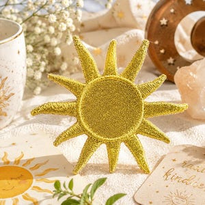 Könnte beinhalten: Ein goldener, gestickter Sonnenaufnäher mit strahlenden Strahlen. Die Sonne ist der Mittelpunkt, umgeben von einer weißen Keramiktasse mit einem Sonnendesign, einem dekorativen Mond und einer Karte mit der Aufschrift "radiate kindness".
