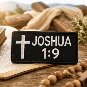 May include: A rectangular black embroidered patch featuring a white cross on the left side and the white text JOSHUA 1:9 on the right side. The patch has a stitched border and is designed with a clean, high-contrast aesthetic for clear visibility.