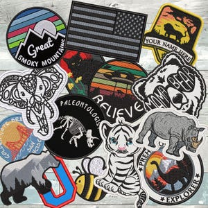 Puede incluir: Una colecci&oacute;n de parches bordados con diversos dise&ntilde;os, incluyendo un paisaje de las Great Smoky Mountains, una bandera estadounidense en blanco y negro, un le&oacute;n, un elefante, un gato negro, un OVNI, un esqueleto de dinosaurio, un oso, una abeja, un tigre y un rinoceronte.