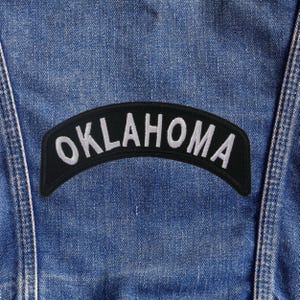 May include: A black, arched patch with the word "OKLAHOMA" embroidered in white. The patch is affixed to a blue denim fabric, likely a jacket or vest. The stitching is visible, and the overall aesthetic is casual and vintage.