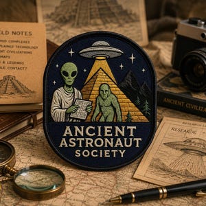 Op de afbeelding: Een ronde geborduurde patch met een donkerblauwe achtergrond met een groene alien die een boek vasthoudt, een groen wezen, een gele piramide en een UFO die een lichtstraal uitstraalt. De tekst ANCIENT ASTRONAUT SOCIETY is onderaan in wit geborduurd.