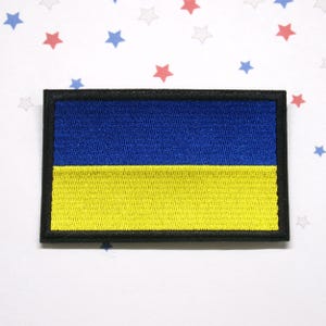 May include: Embroidered patch featuring the flag of Ukraine. The rectangular patch has a blue upper half and a yellow lower half, with a black border. The background is white with red, blue, and gray stars.