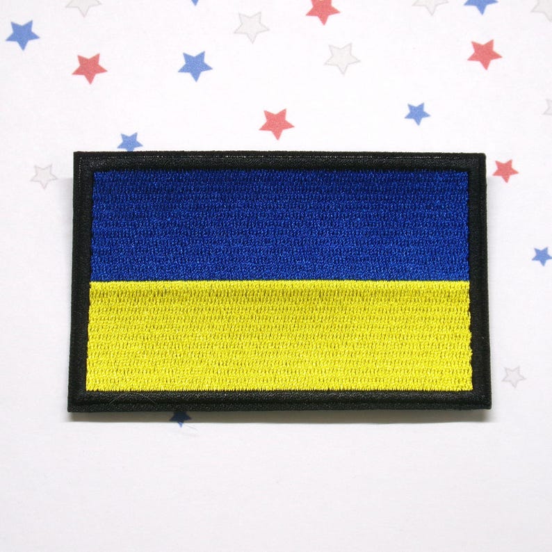 May include: Embroidered patch featuring the flag of Ukraine. The rectangular patch has a blue upper half and a yellow lower half, with a black border. The background is white with red, blue, and gray stars.