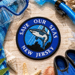 May include: A circular embroidered patch featuring a blue background with a shark illustration and the white text "SAVE OUR SEAS NEW JERSEY" along the border. The patch is surrounded by snorkelling gear, a blue towel, and seashells.