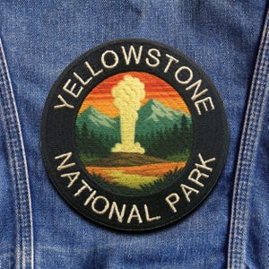May include: A circular embroidered patch featuring the text "YELLOWSTONE NATIONAL PARK" in white lettering. The patch depicts a geyser erupting with a sunset backdrop, mountains, and trees. The patch is sewn onto a denim jacket.