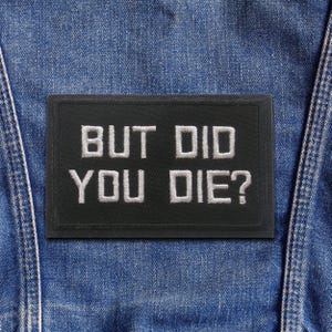 May include: A rectangular black patch with white text that reads "BUT DID YOU DIE?" is sewn onto a blue denim garment. The patch has a white border and is centered on the denim.