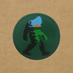 Peut inclure: Un écusson rond et vert brodé représentant une silhouette noire de Bigfoot en marche. Le motif à l'intérieur de la silhouette comprend un ciel bleu, un soleil orange et des arbres verts. Le patch est sur fond beige.