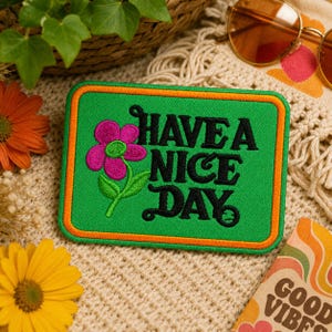 May include: Green rectangular patch with orange trim and the words "HAVE A NICE DAY" in black script. A pink flower with green leaves is embroidered on the patch. The patch is surrounded by flowers and sunglasses.