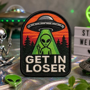 Op de afbeelding: Een rechthoekige geborduurde patch met afgeronde bovenhoeken met een groene alien onder een zilveren UFO die een groene lichtstraal uitstraalt tegen een oranje en rode zonsonderganghemel met zwarte dennenboom-silhouetten en de tekst GET IN LOSER in het wit.