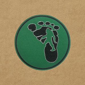 Peut inclure: Un écusson vert circulaire avec un motif brodé noir et blanc. Le motif représente une empreinte de pas et la silhouette d'une personne en randonnée. L'écusson est sur un fond de tissu beige.