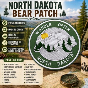 May include: A circular embroidered patch featuring a green border with the text WANDER OFTEN and NORTH DAKOTA. The center depicts a white bear silhouette filled with mountain and pine tree line art and a yellow sun against a green background.