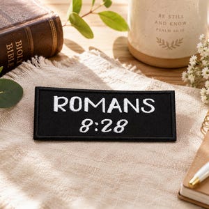 May include: Black rectangular patch with the text "ROMANS 8:28" embroidered in white. The patch is on a beige cloth, with a Bible, mug, notebook, and pen in the background. The mug has the text "BE STILL AND KNOW PSALM 46:10".