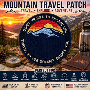 May include: A circular embroidered patch featuring a dark blue background with a yellow sun rising over light blue mountain peaks. The text reads: DON'T TRAVEL TO ESCAPE LIFE, TRAVEL SO LIFE DOESN'T ESCAPE YOU. The patch has a dark blue border.