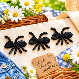 May include: A cream-colored tote bag with three black ant appliqués. The bag is placed on a blue and white checkered cloth, surrounded by daisies, yellow flowers, and a woven basket. A small tag reads "little things matter."