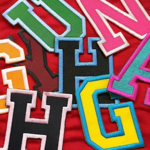 May include: Close-up of colorful embroidered fabric letters in various colors, including green, pink, yellow, and black, against a red background. The letters have a varsity style with contrasting borders, creating a layered effect. Ideal for crafting or personalization projects.
