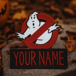 Puede incluir: Parche bordado con el logotipo de Cazafantasmas, un fantasma blanco dentro de un círculo rojo con una línea roja diagonal. Debajo, un parche negro con el texto "YOUR NAME" en rojo. Ideal para Halloween.