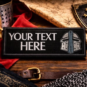 May include: Black rectangular patch with the text "YOUR TEXT HERE" in white, and a helmet design. The patch is surrounded by medieval-themed items, including a shield, chainmail, and a map, all on a wooden surface.