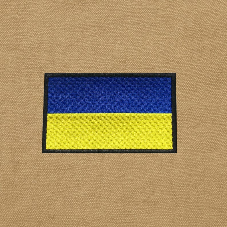 May include: Embroidered patch featuring the Ukrainian flag. The rectangular patch has a blue upper half and a yellow lower half, outlined with a black border. The patch is set against a tan fabric background.