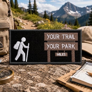 May include: A rectangular black embroidered patch featuring a white stick figure hiker with a backpack and walking stick on the left. On the right, brown wooden-style signs display the white text YOUR TRAIL, YOUR PARK, and MILES.
