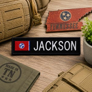 Puede incluir: Un parche negro bordado con el nombre "JACKSON" en blanco. Presenta la bandera del estado de Tennessee en el lado izquierdo. También se ven un recorte de madera con forma de Tennessee y un parche "MADE IN TN".